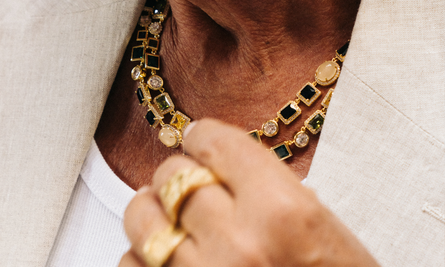Close-up of a layered gold necklace featuring square black stones and round gemstones, worn with a white vest and beige blazer. A gold ringed hand is slightly blurred in the foreground, adjusting the collar.