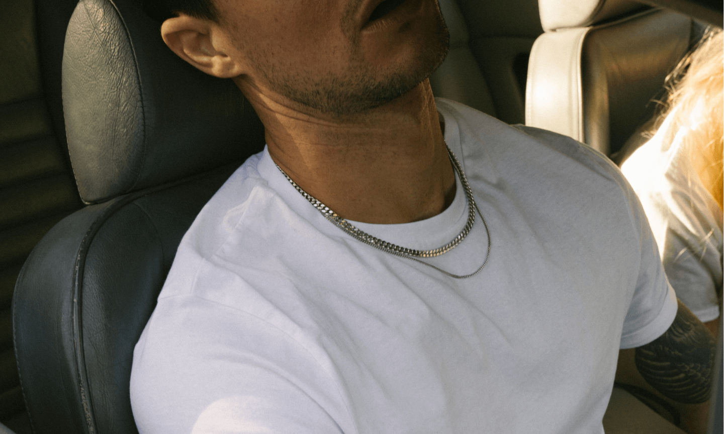Man wearing layered 925 Sterling Silver Cuban Link Chains while lounging in the front seat of a car, styled casually with a white T-shirt under natural sunlight
