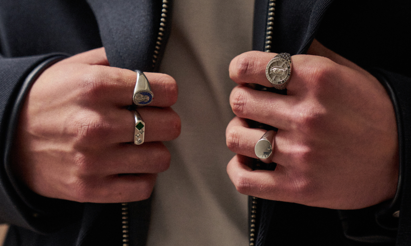 Pair of man's hands gripping the edges of a jacket, each stacked with bold silver rings such as, enamel, gemstones, and minimalist signets.