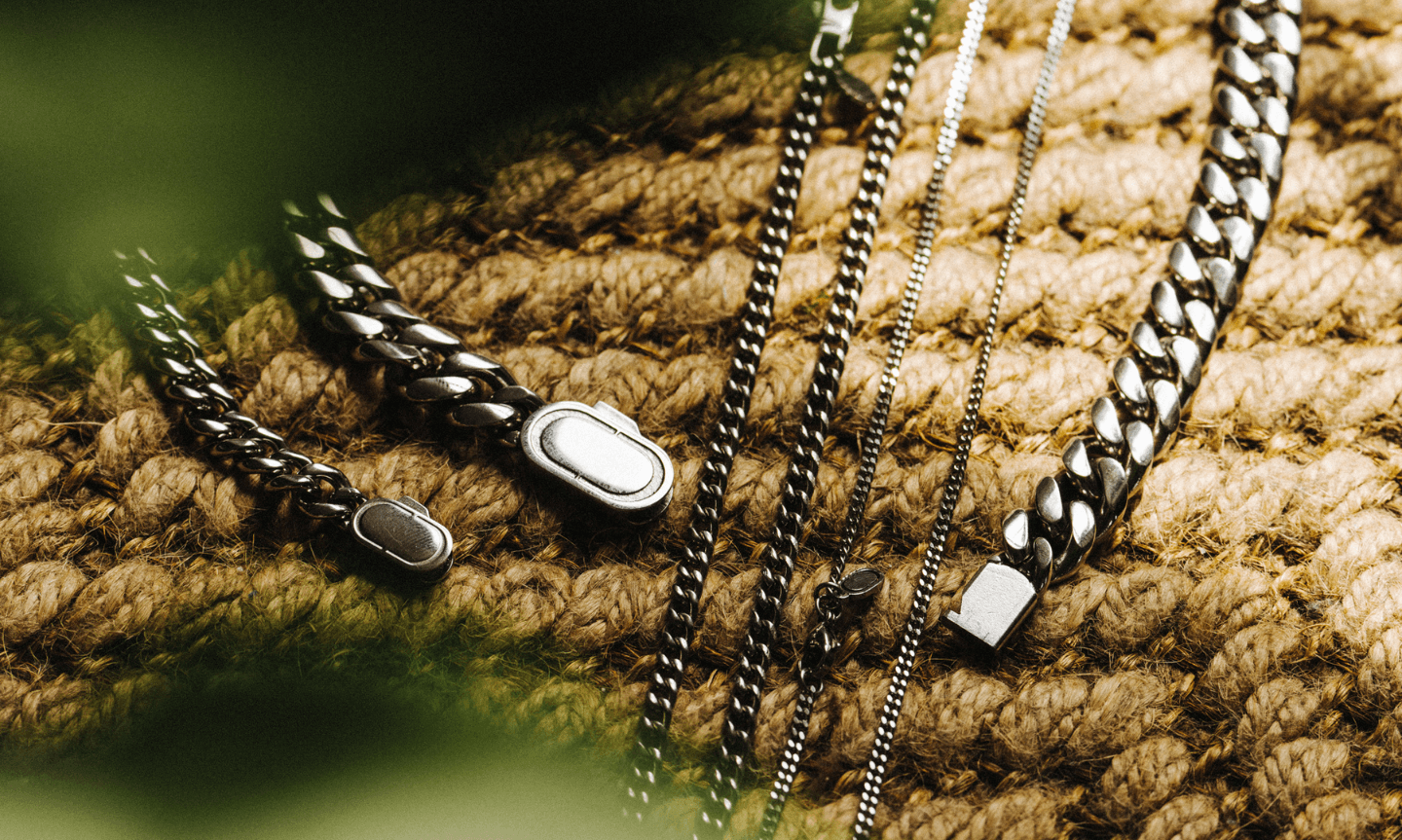 Assorted Sterling Silver chains and bracelets, including Cuban and box link styles, arranged on a woven surface with soft natural light and greenery in the foreground.