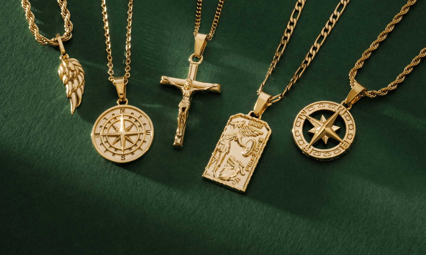 Flat lay of five gold CRAFTD pendants necklaces set against a dark green background. Designs include faith and religious pendants such as an angel wing, crucifix and Saint Michael.